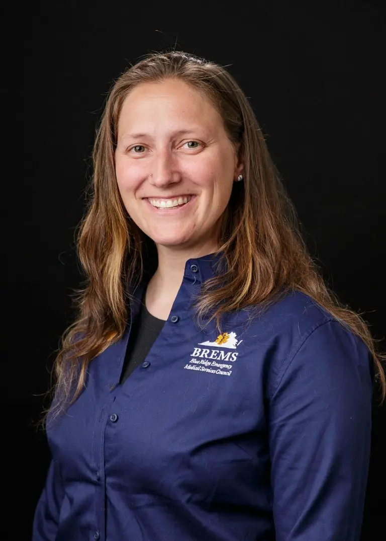 A woman with long brown hair is smiling at the camera. She is wearing a blue button-down shirt with an embroidered logo and text reading "BREMS," "Blue Ridge Emergency Medical Services Council." The background is black.