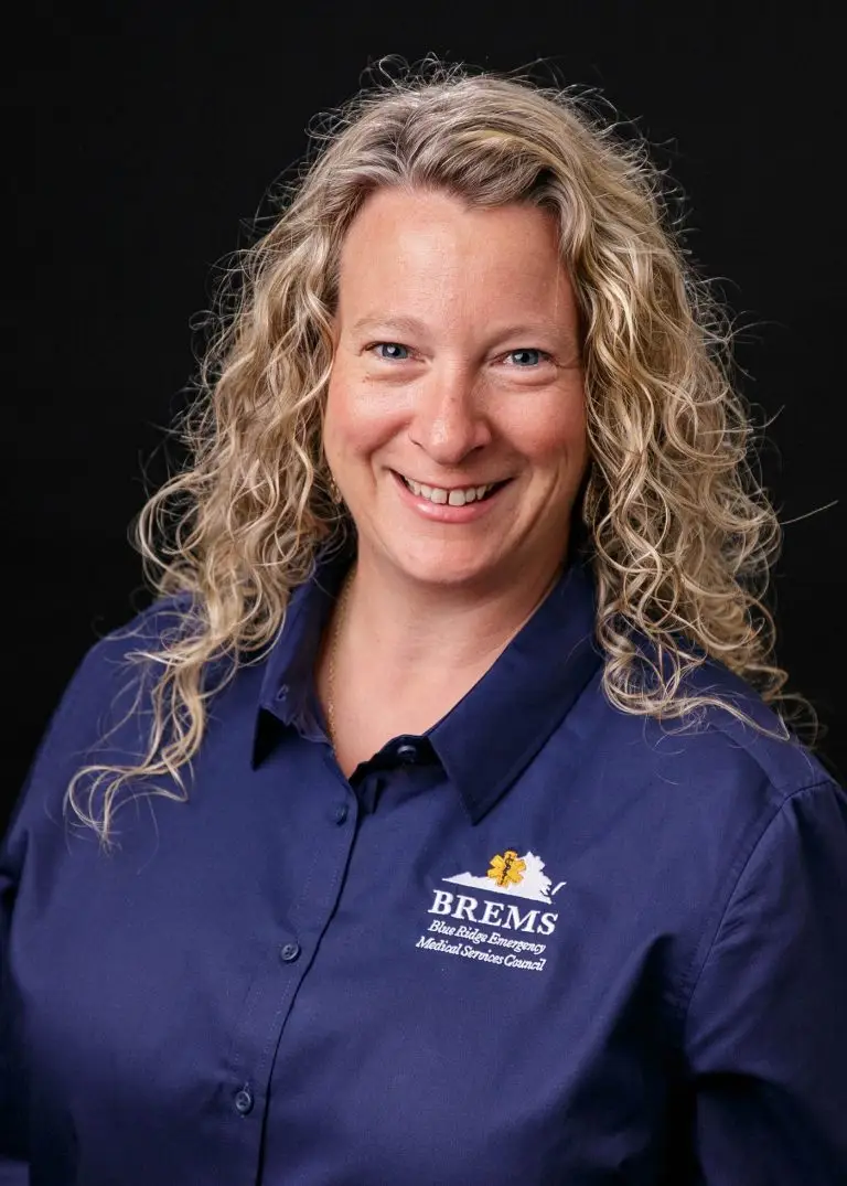 A woman with curly blonde hair smiles while wearing a navy blue shirt with "BREMS Blue Ridge Emergency Medical Services Council" logo embroidered on the chest. She stands against a black background.