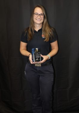 A woman with long hair and glasses, wearing a black polo shirt and dark pants, stands in front of a dark backdrop. She is smiling and holding a shiny award plaque with both hands.