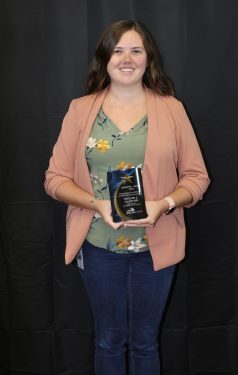A person with long hair stands in front of a dark background, holding a clear award. They are wearing a light pink blazer over a green floral blouse and blue jeans, and they are smiling at the camera.