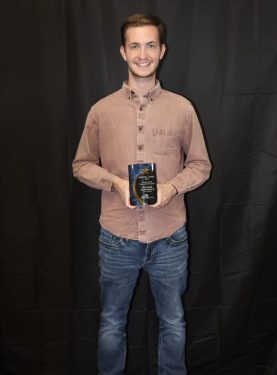 A smiling person stands in front of a dark curtain holding a trophy or award in both hands. They are wearing a brown button-up shirt and blue jeans.