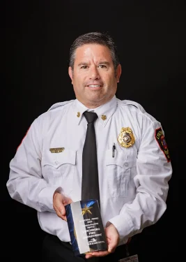 A man in uniform is holding a trophy. He is wearing a white shirt with badges, a black tie, and has a fire department patch on his sleeve. The trophy he holds reads "National Award for the Firefighting Department." The background is plain and dark.