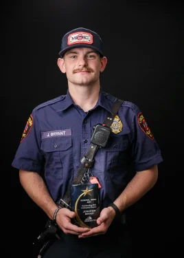 A firefighter in a navy uniform and cap, labeled "J. Bryant" and "MEDIC," stands against a black background, holding an award plaque. The plaque has text visible but not readable in the image. The firefighter has a mustache and a calm expression.