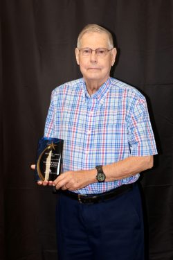 An elderly man wearing glasses and a plaid shirt stands against a dark background holding a plaque, which appears to be an award, in his right hand. The man has short, white hair and is looking directly at the camera.