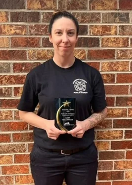 A person with dark hair pulled back stands in front of a brick wall holding an award. They are wearing a black t-shirt, which has a badge logo on the left side with the text "Public Safety.