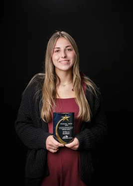 A woman with long blonde hair wearing a red dress and black cardigan stands against a dark background, smiling and holding a plaque or award with both hands. The award has a star design on it.